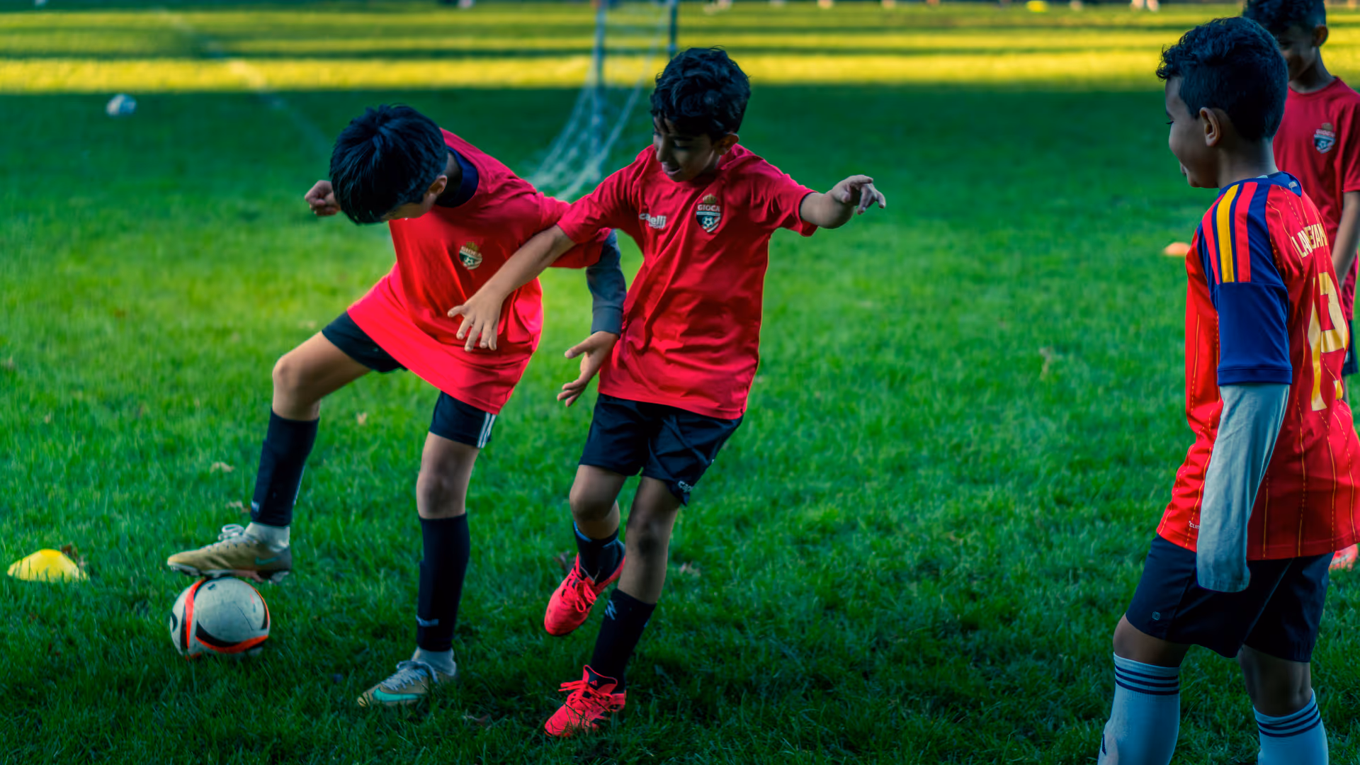 Two Gioca players contesting the ball in a sunlit one-against-one drill as a teammate watches
